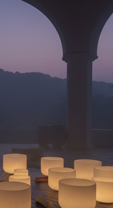 Cinematic dark editorial photograph of a serene sound bath environment at dusk — a set of crystal singing bowls and a large hammered metal gong glowing softly in warm champagne-gold light, deep espresso shadows, atmospheric haze, luxurious five-star wellness resort mood. Moody, sophisticated, high contrast, spacious negative space. No people, no text.